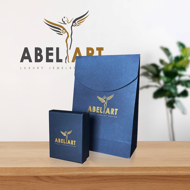 May include: Navy blue jewelry box and gift bag with gold foil logo of a winged figure and the text "ABEL ART LUXURY JEWELRY DESIGN". The packaging sits on a wooden surface, with a logo in the background.