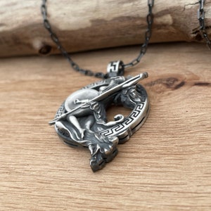 May include: A silver pendant necklace featuring a detailed depiction of a classical figure with a shield and spear. The pendant is circular with a Greek key design around the edge and hangs from a dark chain. The background is a wooden surface.
