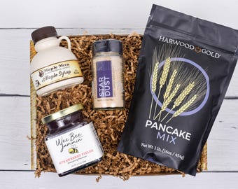 May include: A gift basket filled with breakfast items. It includes a bottle of maple syrup, a jar of strawberry preserves, a spice blend, and a bag of pancake mix. The pancake mix is 1 lb (454g).