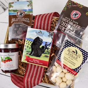 May include: A gift basket filled with cherry-themed treats. It includes a jar of cherry preserves, bags of milk chocolate-covered cherries, milk and dark chocolate cherry turtles, and snow-covered dried cherries. A red and white striped ribbon accents the basket.