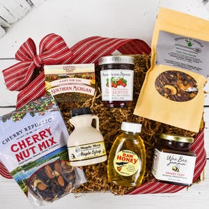 May include: A gift basket filled with gourmet food items. It includes a bag of Cherry Nut Mix, a jar of strawberry preserves, a bottle of raw honey, a jar of sweet tart jam, a bag of granola, and a small maple syrup jug. A red and white striped bow adorns the basket.