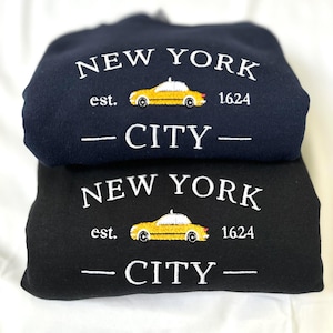 以下が含まれることがあります： 白い刺繍が施されたネイビーブルーとブラックの2つのスウェットシャツ。スウェットシャツには、「NEW YORK CITY」の文字と、黄色のタクシーのグラフィック、そして「est. 1624」の文字が描かれています。