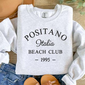 May include: Gray sweatshirt with black text that reads "Positano Italia Beach Club - 1995 -"