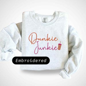 Könnte beinhalten: Hellgraues Sweatshirt mit dem gestickten Schriftzug "Dunkie Junkie" in orange und pinkfarbener Schrift, begleitet von einer Kaffeetassen-Grafik. Das Wort "Embroidered" befindet sich auf einem schwarzen Oval.