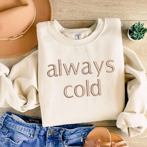 Puede incluir: Una sudadera de color crema con el texto "always cold" en letras marrones.