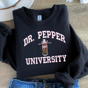 May include: Black sweatshirt with a pink graphic of a Dr. Pepper bottle and the text "Dr. Pepper University".