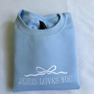 May include: A light blue sweatshirt with the text "JESUS LOVES YOU" embroidered in white. The text is accompanied by a white embroidered bow.