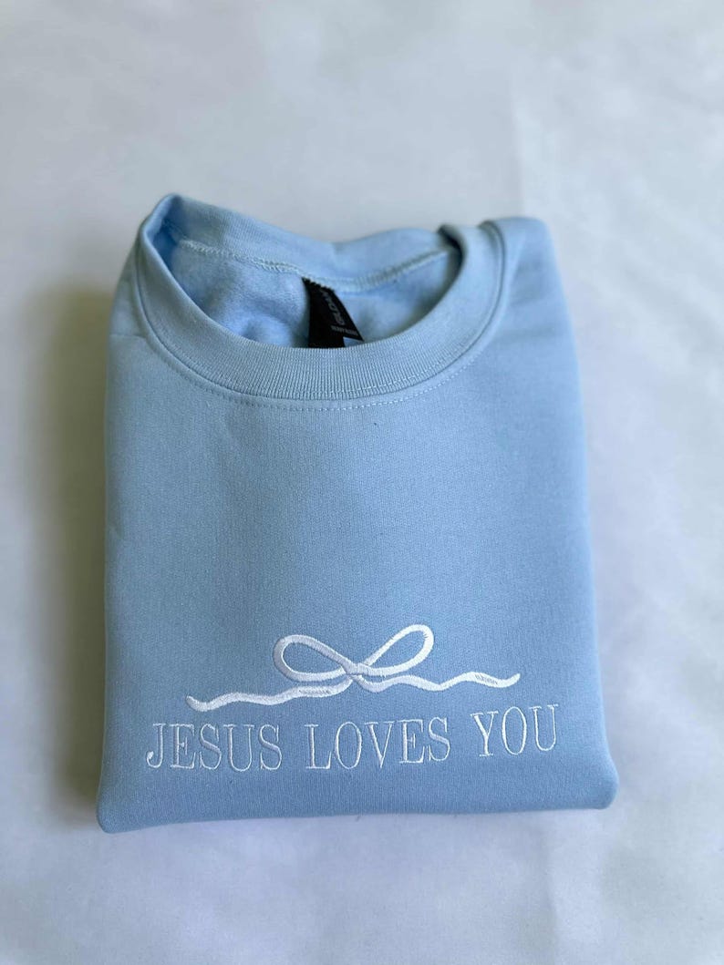 May include: A light blue sweatshirt with the text "JESUS LOVES YOU" embroidered in white. The text is accompanied by a white embroidered bow.