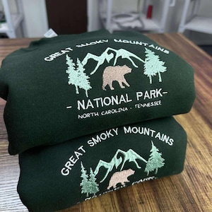 Könnte beinhalten: Zwei dunkelgrüne Sweatshirts sind gestapelt, jedes mit "GREAT SMOKY MOUNTAINS NATIONAL PARK" bestickt. Das Design umfasst Berge, Bäume und einen Bären. Der Text enthält auch "NORTH CAROLINA . TENNESSEE."