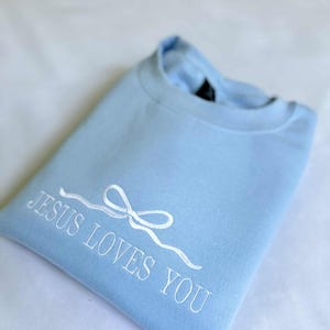 May include: A light blue sweatshirt with the text "JESUS LOVES YOU" embroidered in white. A white ribbon bow is above the text.