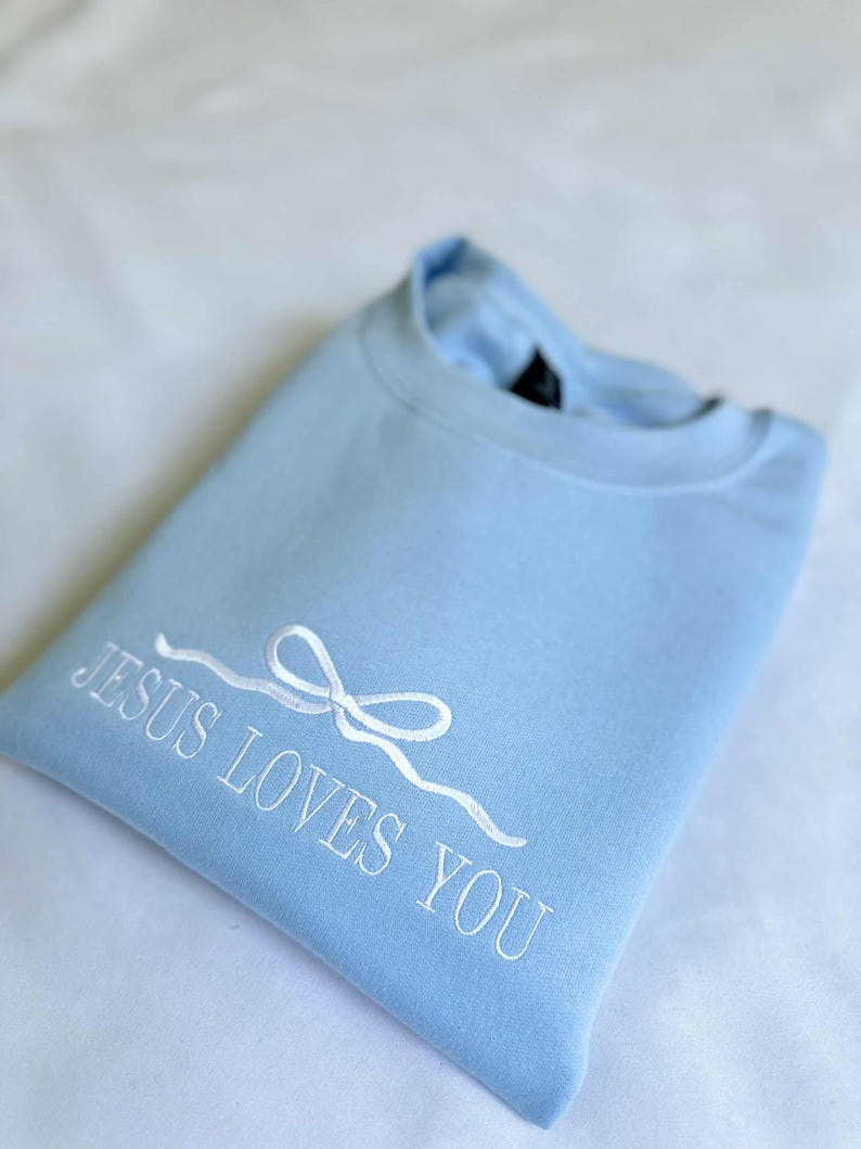 May include: A light blue sweatshirt with the text "JESUS LOVES YOU" embroidered in white. A white ribbon bow is above the text.