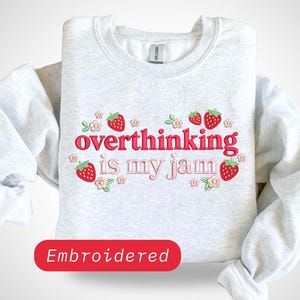 May include: A light gray crewneck sweatshirt featuring embroidered red strawberries and small pink flowers surrounding the text "overthinking is my jam" in pink and red lettering. The design is centered on the chest of the long-sleeved garment.