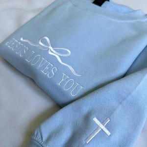 May include: Light blue sweatshirt with white embroidery that reads "Jesus Loves You" and a white cross on the sleeve.