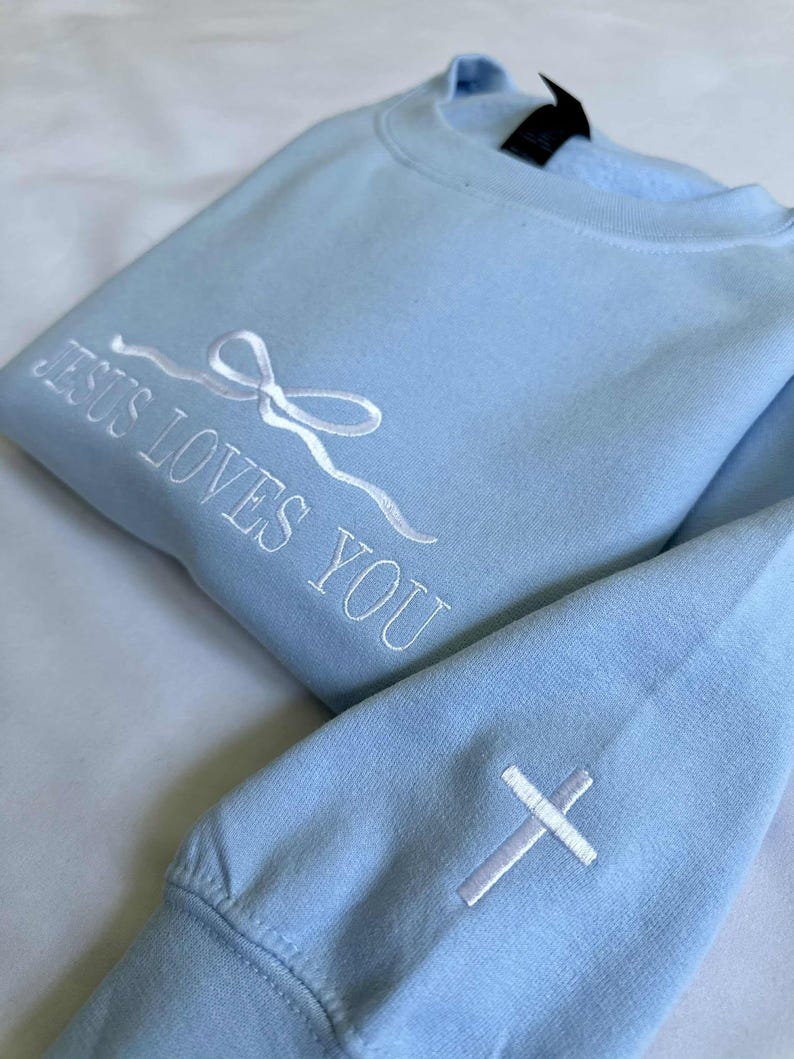 May include: Light blue sweatshirt with white embroidery that reads "Jesus Loves You" and a white cross on the sleeve.