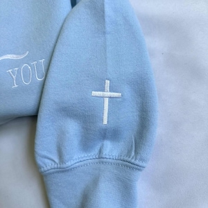 May include: A light blue sweatshirt with a white embroidered cross on the sleeve and the word "YOU" on the front.