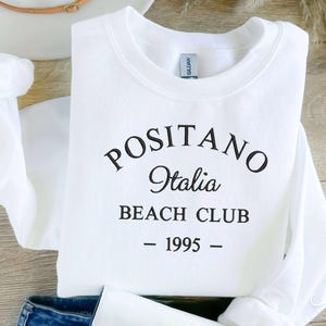 May include: White sweatshirt with black text that reads "Positano Italia Beach Club - 1995 -"