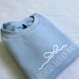 May include: A light blue sweatshirt with a white embroidered design that says "JESUS LOVES YOU" with a stylized white bow.