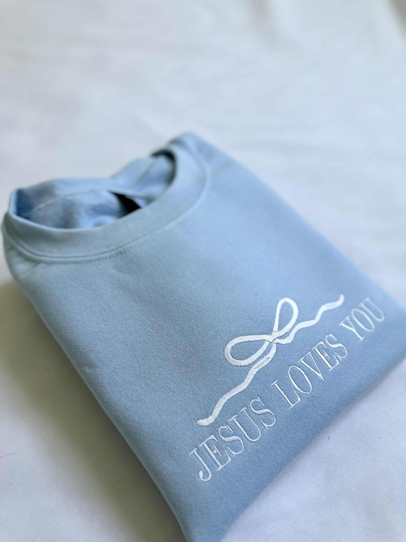 May include: A light blue sweatshirt with a white embroidered design that says "JESUS LOVES YOU" with a stylized white bow.