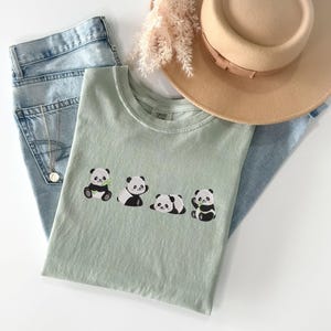 May include: A light green t-shirt with four embroidered panda bears. The pandas are black and white and are holding bamboo shoots. The t-shirt is laying on a pair of blue jeans and a straw hat.