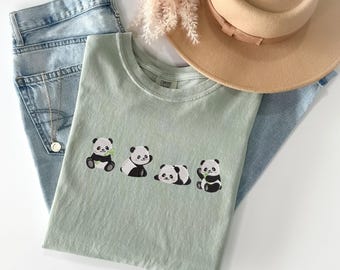 Op de afbeelding: Een lichtgroen T-shirt met vier geborduurde pandaberen. De panda's zijn zwart-wit en houden bamboescheuten vast. Het T-shirt ligt op een blauwe spijkerbroek en een strohoed.