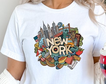 May include: White t-shirt featuring a colourful doodle design of New York City landmarks and icons. The design includes the Statue of Liberty, the Empire State Building, the Brooklyn Bridge, and a yellow taxi cab. The words "New York" are written in large, bold letters.