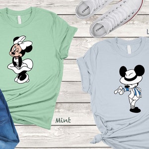 Può includere: Due t-shirt con un'immagine di cartone animato di Topolino e Minnie Mouse. La maglietta di Topolino è blu chiaro e la maglietta di Minnie Mouse è verde menta. Il testo "Light Blue" e "Mint" è stampato sotto le magliette.