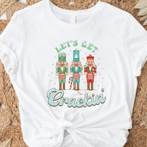 May include: White t-shirt with a Christmas-themed design featuring three nutcracker figures playing drums and the text "Let's Get Crackin'".