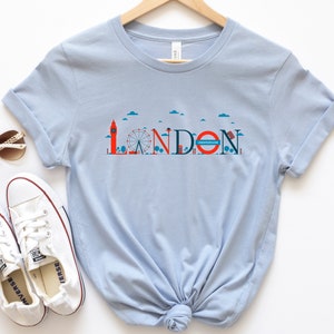 May include: Light blue t-shirt featuring a London-themed graphic. The design includes the word "LONDON" with iconic landmarks like Big Ben and the London Eye. The word "UNDERGROUND" is also included in the design.