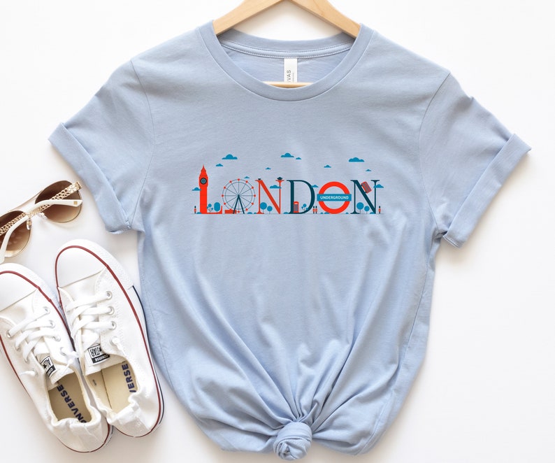 May include: Light blue t-shirt featuring a London-themed graphic. The design includes the word "LONDON" with iconic landmarks like Big Ben and the London Eye. The word "UNDERGROUND" is also included in the design.