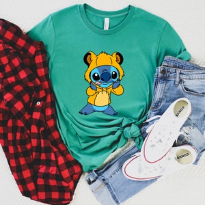 May include: A teal blue t-shirt with a cartoon image of Stitch wearing a yellow lion costume. Stitch is sticking out its tongue and making a silly face.