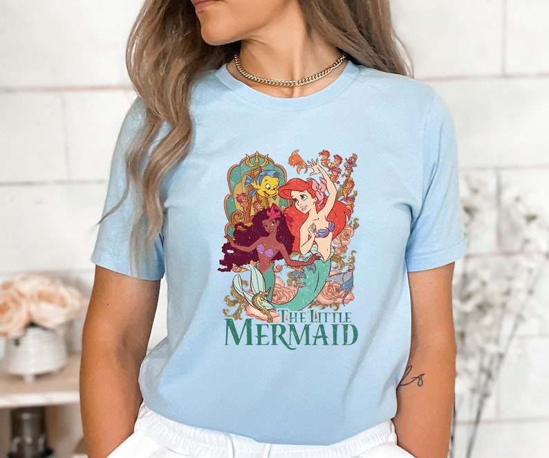 Może przedstawiać: Jasno błękitny t-shirt z nadrukiem graficznym przedstawiającym Ariel, Sebastiana i Floundera z bajki Mała Syrenka. Pod obrazkiem znajduje się napis "The Little Mermaid".