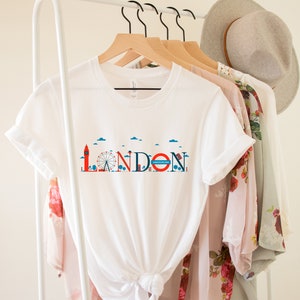 May include: White t-shirt with a London-themed graphic. The design features iconic landmarks like Big Ben and the London Eye, along with the word "LONDON" in a stylized font. The shirt is hanging on a rack with other clothing items.