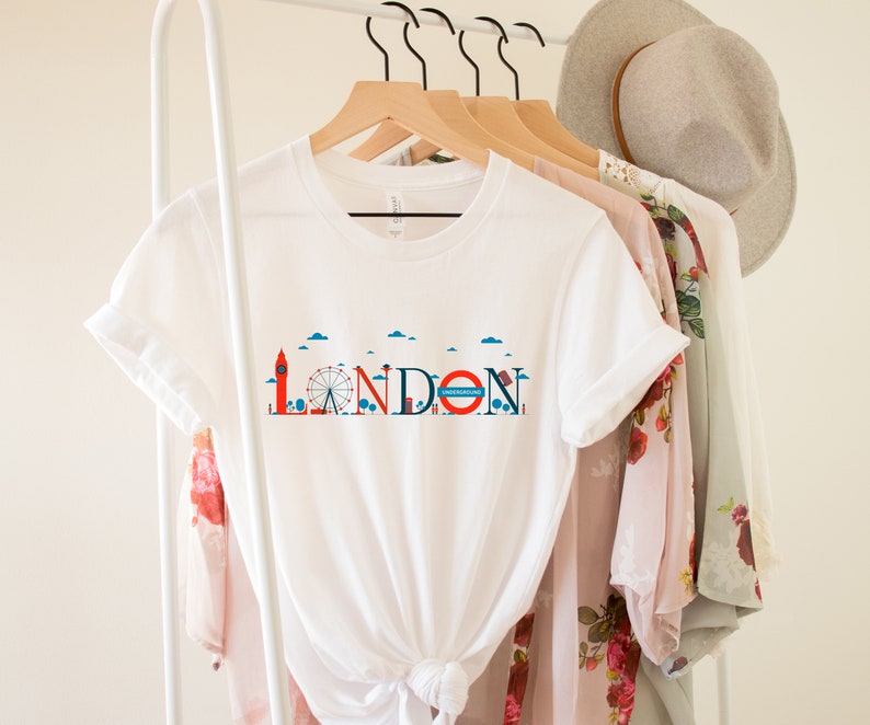 May include: White t-shirt with a London-themed graphic. The design features iconic landmarks like Big Ben and the London Eye, along with the word "LONDON" in a stylized font. The shirt is hanging on a rack with other clothing items.