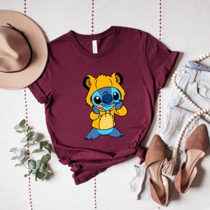 May include: A maroon t-shirt with a cartoon character wearing a yellow hooded sweatshirt. The character has blue fur and big eyes.