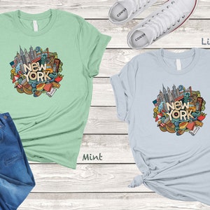 May include: Two t-shirts with a graphic design of a New York City skyline and other iconic symbols. The t-shirt on the left is mint green and the t-shirt on the right is light blue.