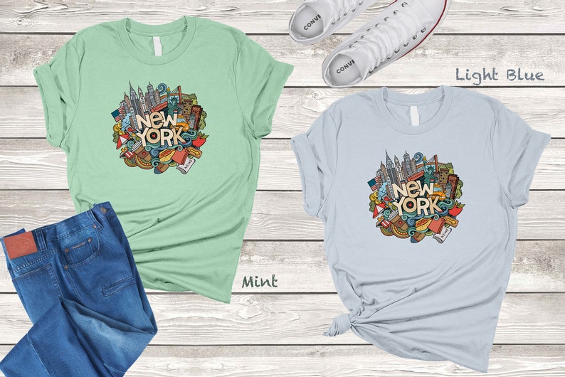 May include: Two t-shirts with a graphic design of a New York City skyline and other iconic symbols. The t-shirt on the left is mint green and the t-shirt on the right is light blue.
