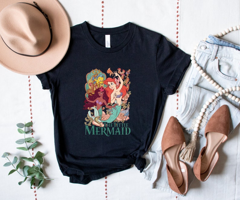 Może przedstawiać: Czarny T-shirt z nadrukiem graficznym przedstawiającym Ariel, Floundera i Urszulę z bajki Mała Syrenka. Pod nadrukiem znajduje się napis "The Little Mermaid".