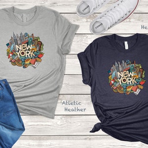 May include: Two t-shirts with a graphic design of New York City landmarks and icons. The shirt on the left is athletic heather and the shirt on the right is heather navy.