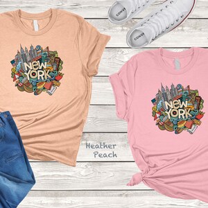 May include: Two t-shirts with a graphic design of a New York City skyline and other iconic symbols. The t-shirt on the left is heather peach and the t-shirt on the right is pink.