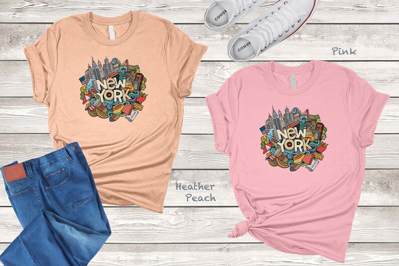 May include: Two t-shirts with a graphic design of a New York City skyline and other iconic symbols. The t-shirt on the left is heather peach and the t-shirt on the right is pink.