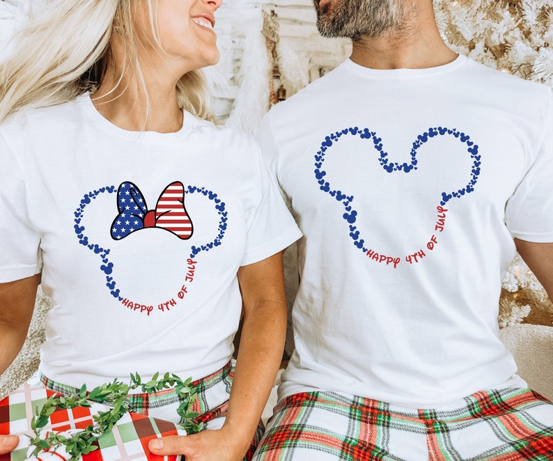 May include: Two white t-shirts with a red, white, and blue American flag bow and Mickey Mouse ears made of blue hearts. The text "Happy 4th of July" is written in red and blue around the ears.