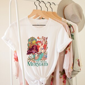 Może przedstawiać: Biała koszulka z grafiką przedstawiającą Ariel, Sebastiana i Floundera z bajki Mała Syrenka. Grafika jest w stylu vintage z kolorowym kwiatowym tłem. Tekst "Mała Syrenka" jest nadrukowany pod grafiką.