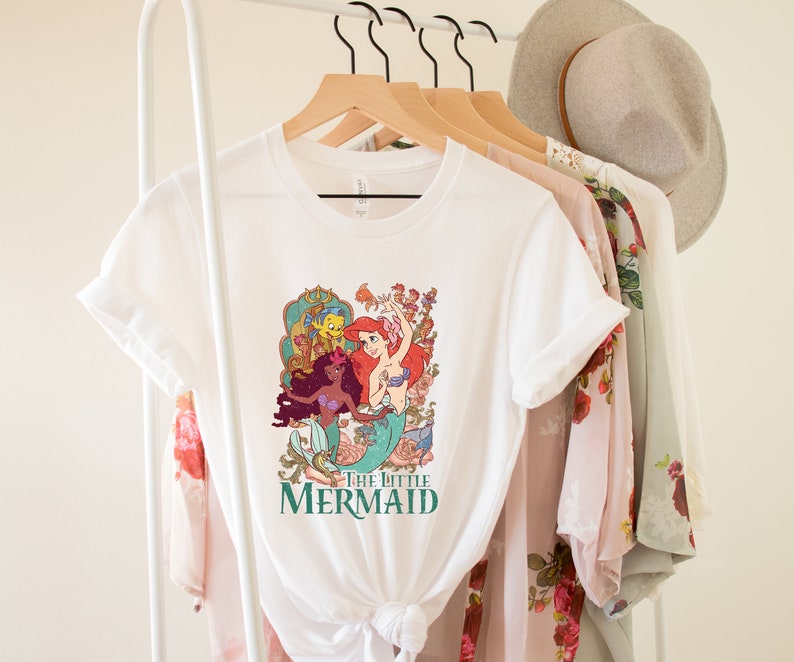 Może przedstawiać: Biała koszulka z grafiką przedstawiającą Ariel, Sebastiana i Floundera z bajki Mała Syrenka. Grafika jest w stylu vintage z kolorowym kwiatowym tłem. Tekst "Mała Syrenka" jest nadrukowany pod grafiką.