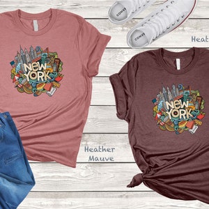 May include: Two t-shirts with a graphic design of a circle of New York City themed doodles, including the Statue of Liberty, the Empire State Building, and the words "New York". The shirt on the left is heather mauve and the shirt on the right is heather maroon.