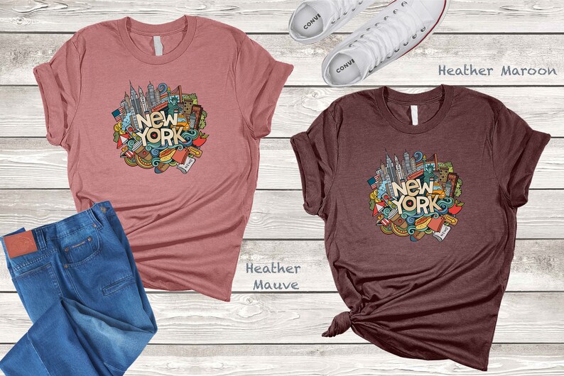 May include: Two t-shirts with a graphic design of a circle of New York City themed doodles, including the Statue of Liberty, the Empire State Building, and the words "New York". The shirt on the left is heather mauve and the shirt on the right is heather maroon.