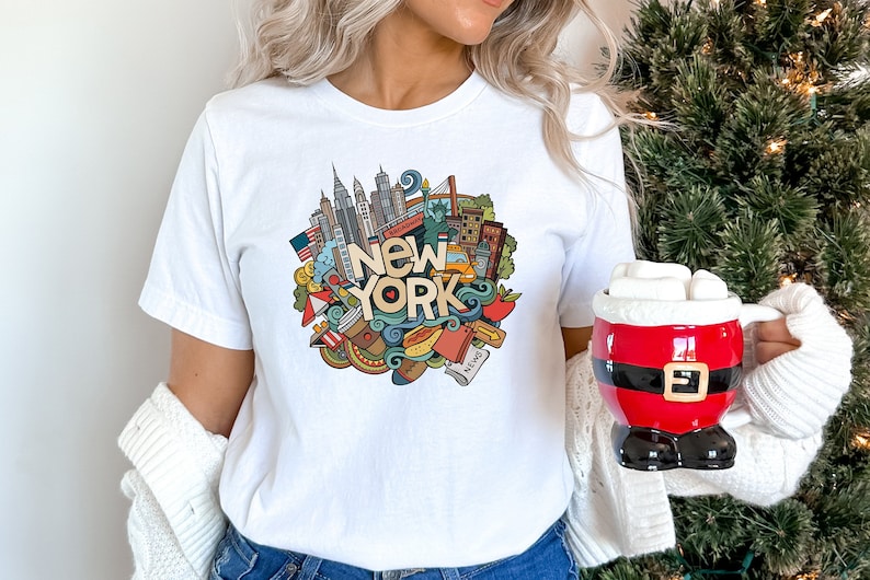 May include: White t-shirt with a colorful doodle design of New York City landmarks and icons. The design includes the Statue of Liberty, the Empire State Building, the Brooklyn Bridge, and a yellow taxi cab. The words "New York" are written in large, bold letters.