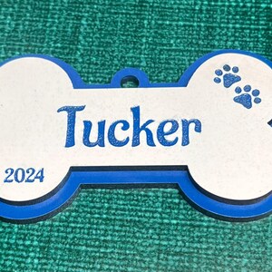Könnte beinhalten: Blau-weißer Knochen-Anhänger mit dem Namen "Tucker" und dem Jahr "2024" darauf. Zwei blaue Pfotenabdrücke befinden sich oben rechts auf dem Anhänger.