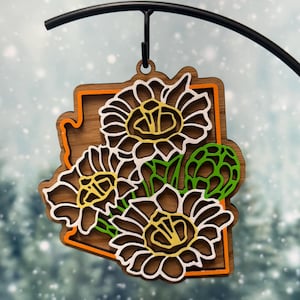 Peut inclure: Ornement en bois représentant des tournesols. L'ornement a la forme de l'État de l'Arizona, avec une base marron, un bord orange et des détails de tournesols blancs, verts et jaunes. Il est suspendu à un crochet en métal noir.