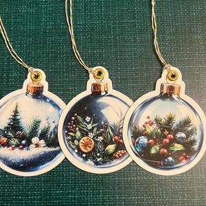 May include: Three Christmas ornament-shaped tags with gold string. Each tag features a different winter scene inside a glass ball. The scenes include a snowy forest, berries, and greenery. The tags have a gold-colored metal cap.