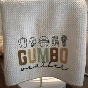 May include: White kitchen towel with a black and white illustration of a pot of gumbo and vegetables. The text "GUMBO weather" is written in brown, yellow, and green letters.